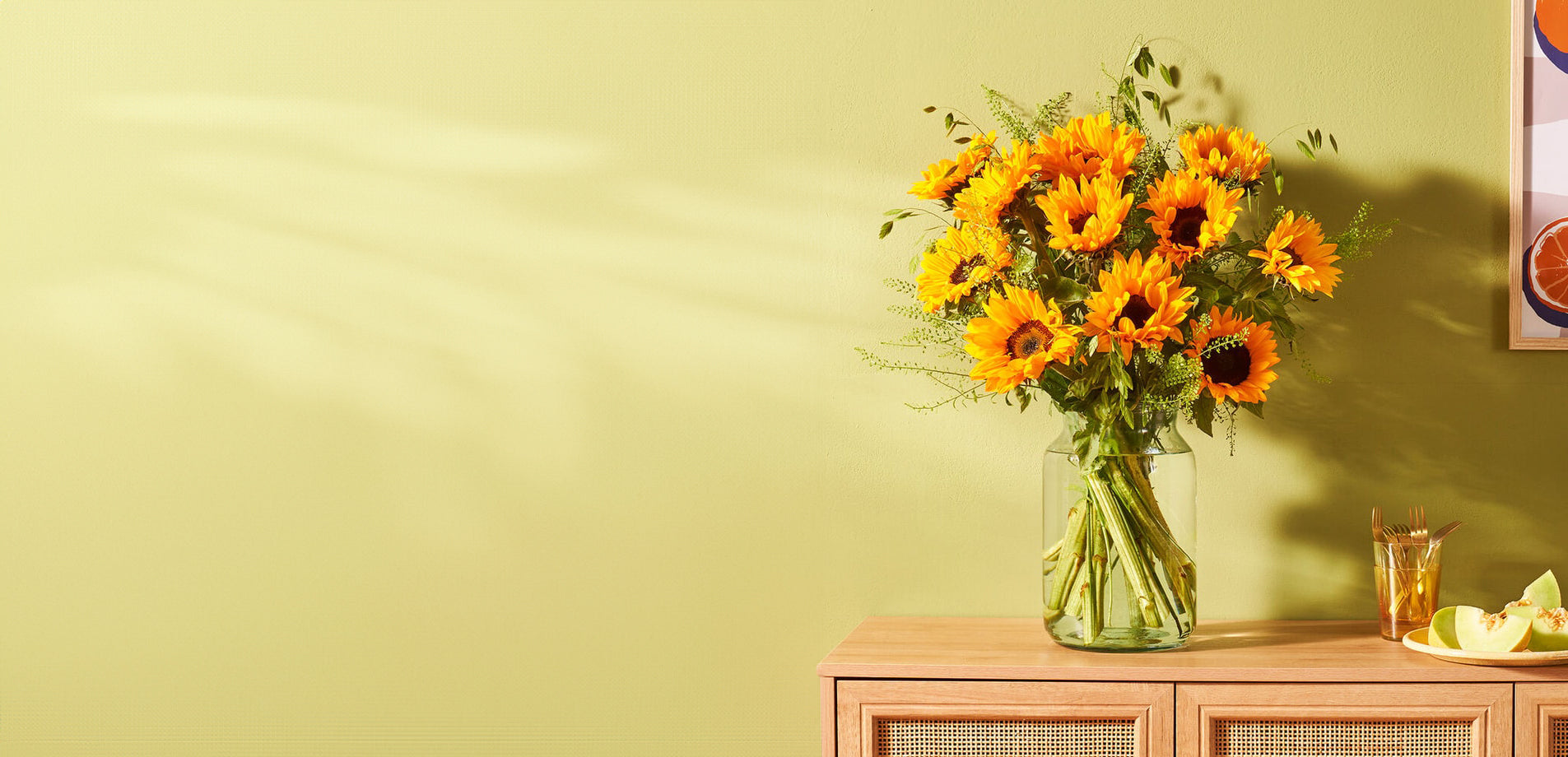 Boeket zonnebloemen in een glazen vaas op een houten kastje tegen een lichtgroene achtergrond.