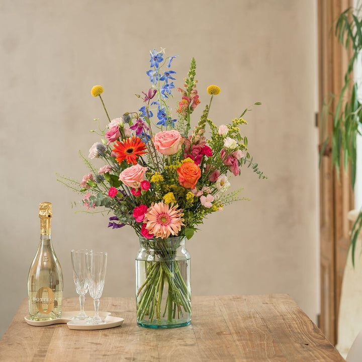 Bont boeket met roze, oranje en blauwe bloemen, waaronder rozen en gerbera’s, in glazen vaas op houten tafel met champagnefles en glazen