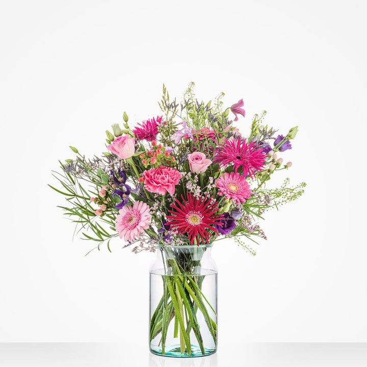 Groot verjaardagboeket in een glazen vaas met roze, paarse en rode bloemen tegen een witte achtergrond.
