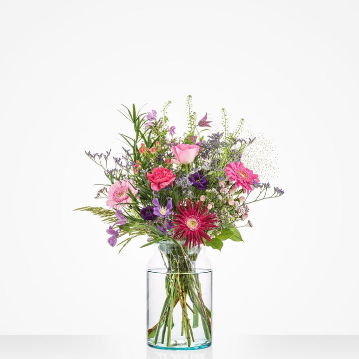 Klein verjaardagboeket in een glazen vaas met roze, paarse en rode bloemen tegen een witte achtergrond.