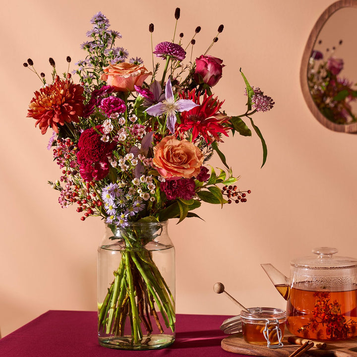 Herfstboeket met rozen, rode dahlia’s en paarse bloemen in glazen vaas, sfeervol gestyled.