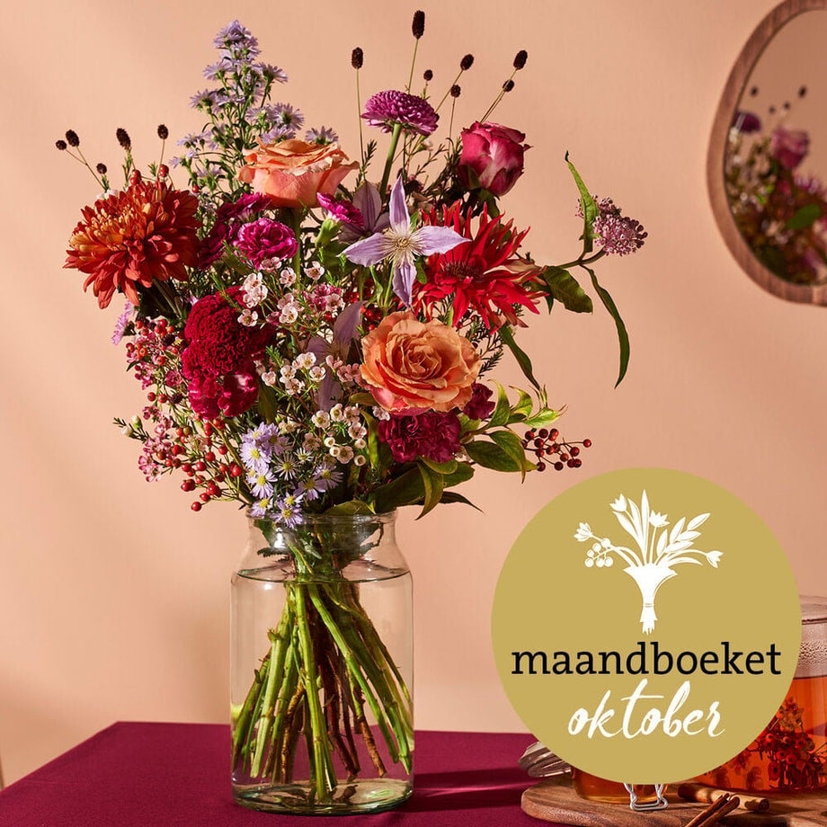 Maandboeket oktober met rozen, dahlia’s en herfstbloemen in een glazen vaas, op een sfeervolle tafel.