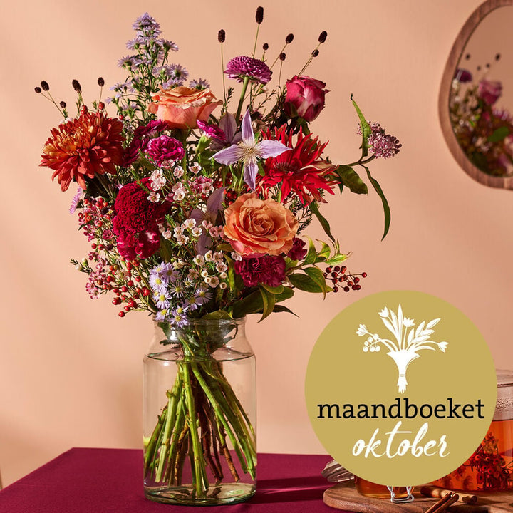 Maandboeket oktober met rozen, dahlia’s en herfstbloemen in een glazen vaas, op een sfeervolle tafel.