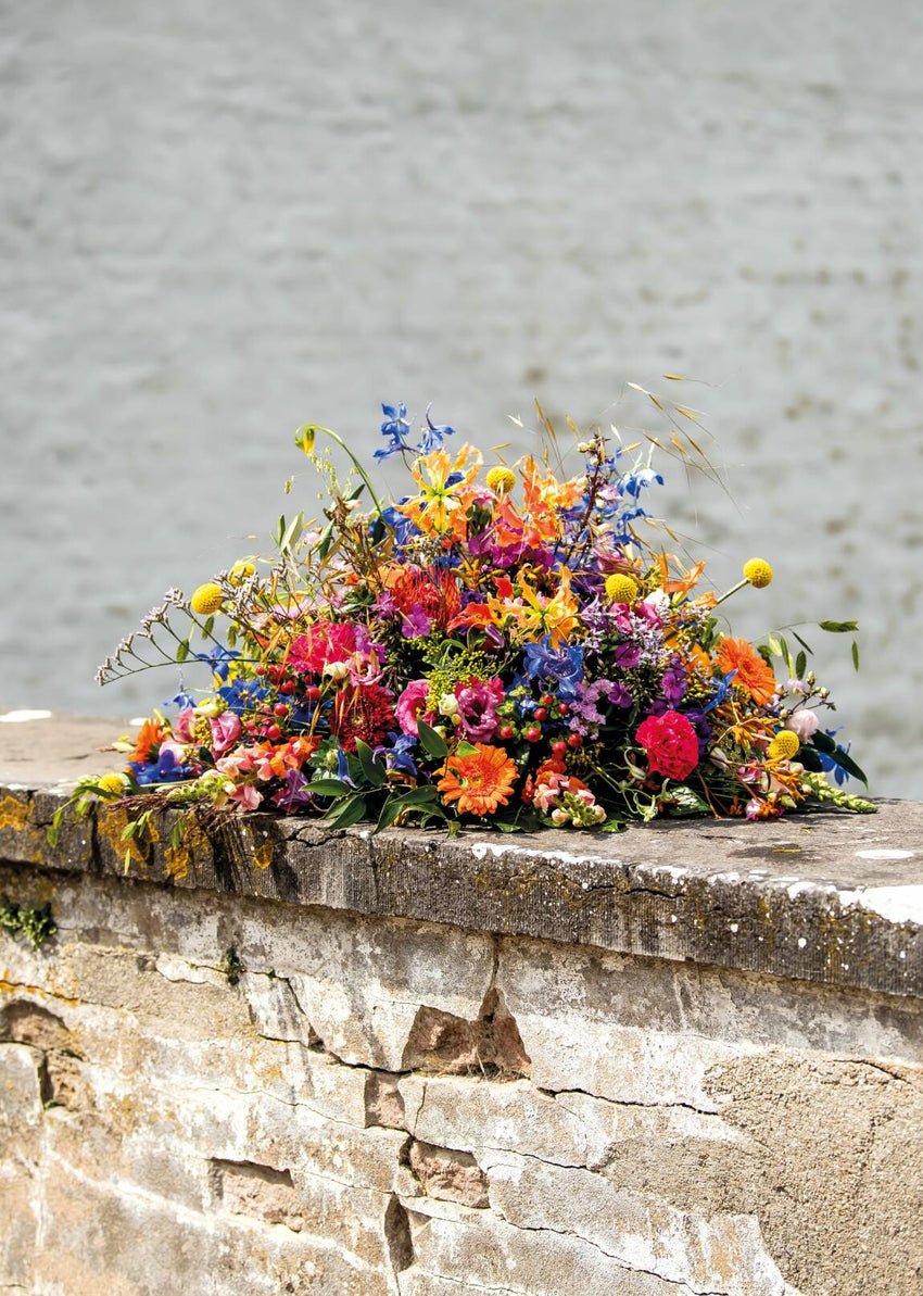 Ovaal rouwstuk met kleurrijke bloemen zoals gerbera’s, rozen en delphinium op stenen muurtje.