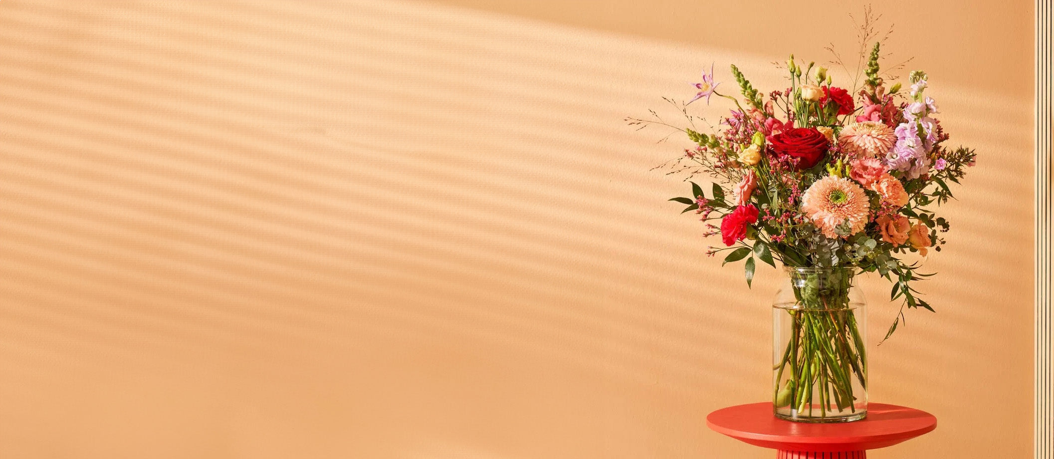 Boeket met anjers en gemengde bloemen in een glazen vaas op een rode tafel tegen een beige achtergrond.