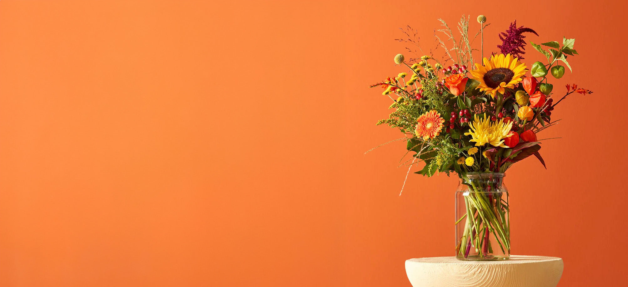 Boeket met chrysanten en gemengde bloemen in een glazen vaas tegen een oranje achtergrond.