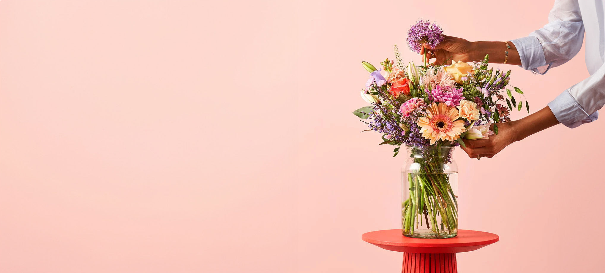 Condoleance boeket met zachte kleuren bloemen in een glazen vaas, geschikt als troostbloemen, op een rode tafel tegen een lichtroze achtergrond.