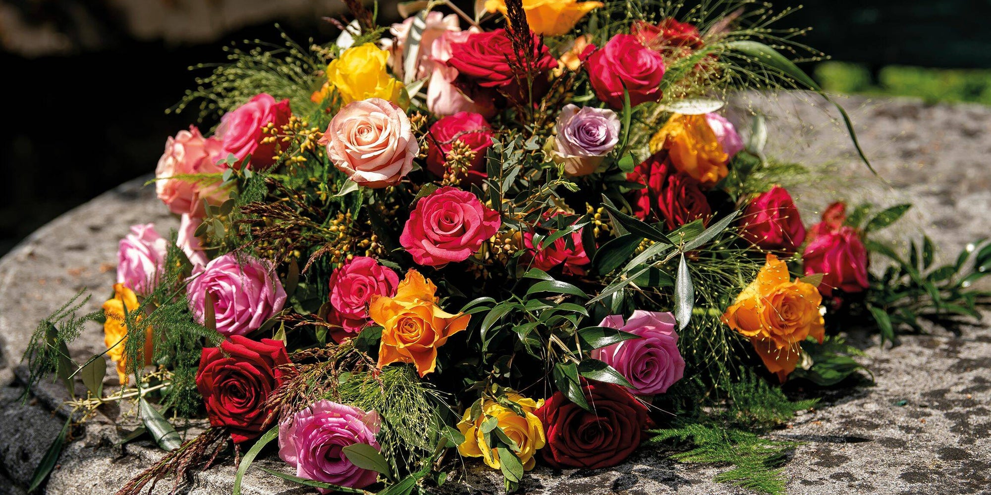 Rouw bloemstuk met kleurrijke rozen in tinten van rood, roze, oranje en paars, geplaatst op een stenen tafel.