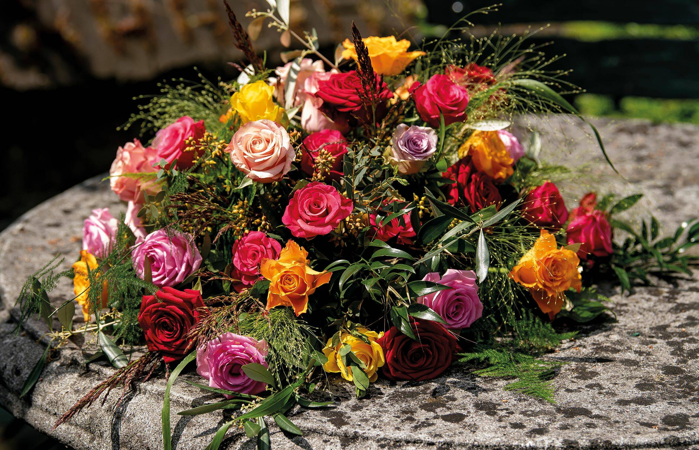 Rouw bloemstuk met kleurrijke rozen in tinten van rood, roze, oranje en paars, geplaatst op een stenen tafel.