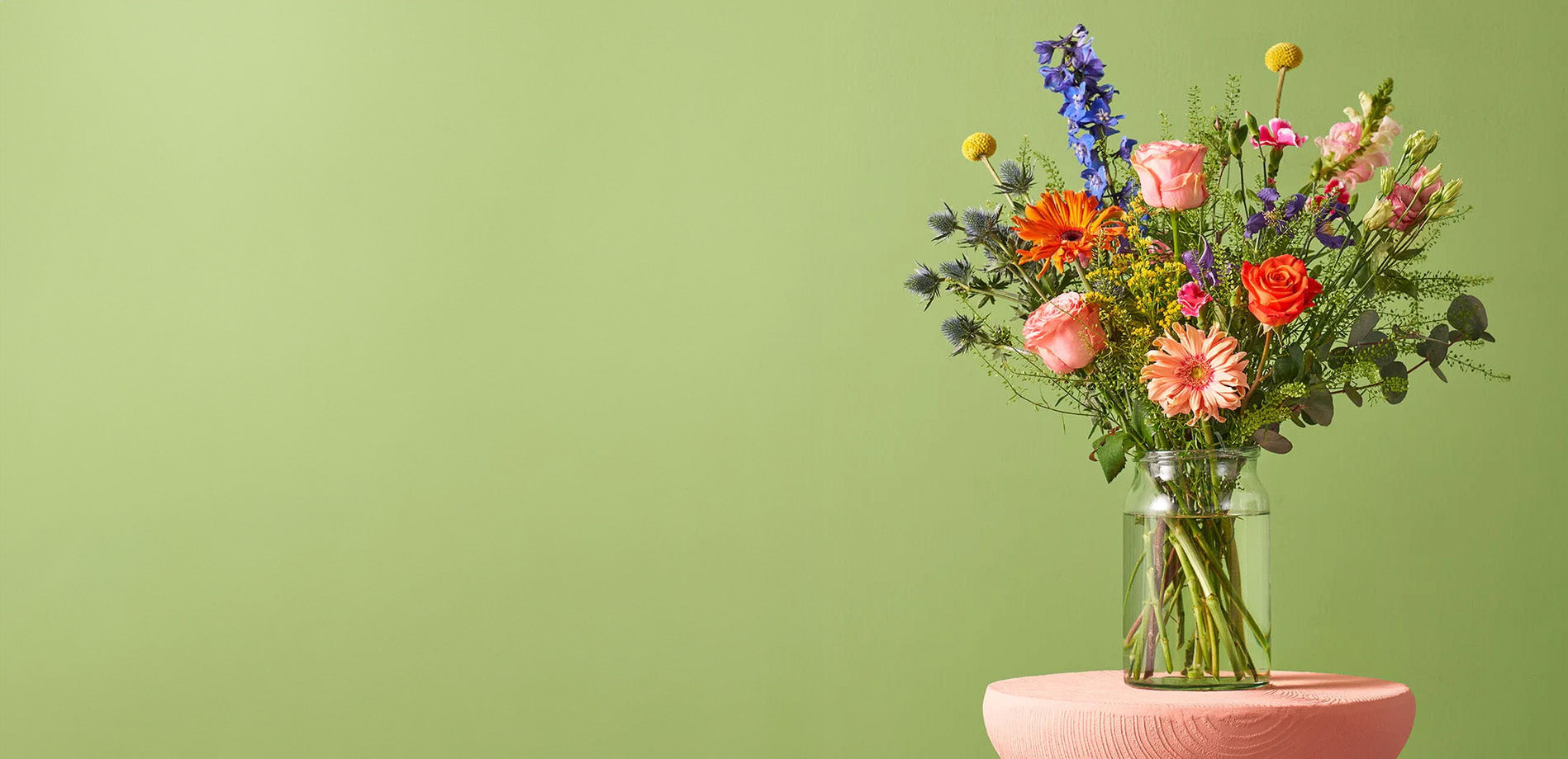 Boeket met gerbera’s en gemengde bloemen in een glazen vaas op een roze tafel tegen een lichtgroene achtergrond.