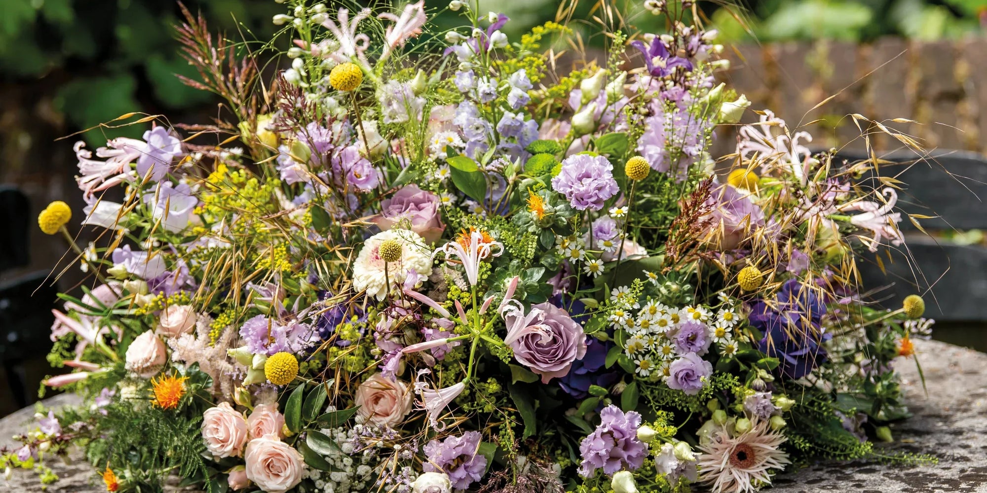 Rouwbloemstuk in zachte tinten van lila, crème en groen, met rozen, gerbera’s, lisianthus en veldbloemen, liggend op een ronde stenen tafel.