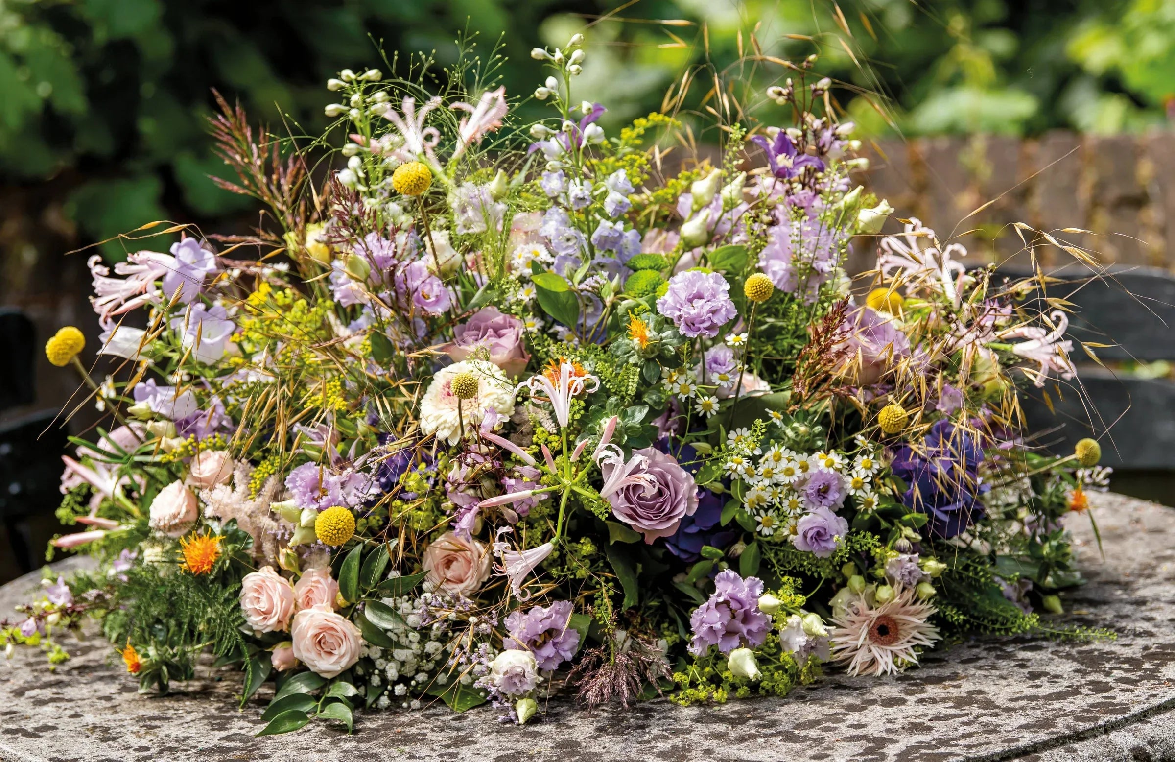 Rouwbloemstuk in zachte tinten van lila, crème en groen, met rozen, gerbera’s, lisianthus en veldbloemen, liggend op een ronde stenen tafel.