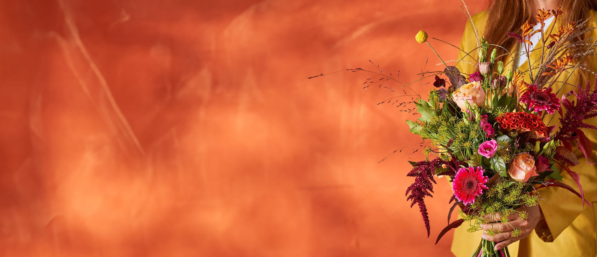 Herfst boeket met kleurrijke bloemen in warme tinten, vastgehouden door een vrouw tegen een oranje achtergrond.