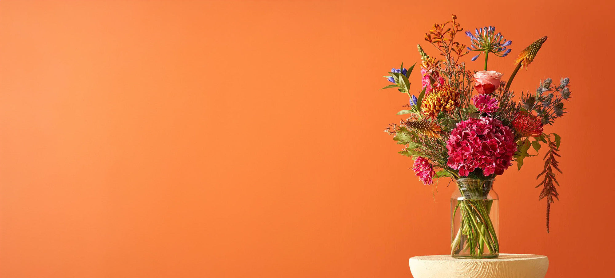 Boeket met hortensia en kleurrijke bloemen in een glazen vaas tegen een oranje achtergrond.