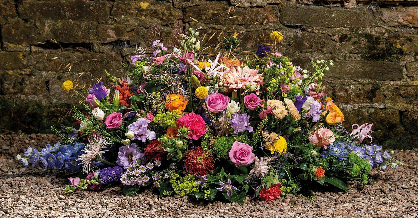Rouwarrangement met een overvloed aan kleurrijke bloemen tegen een natuurlijke stenen achtergrond.