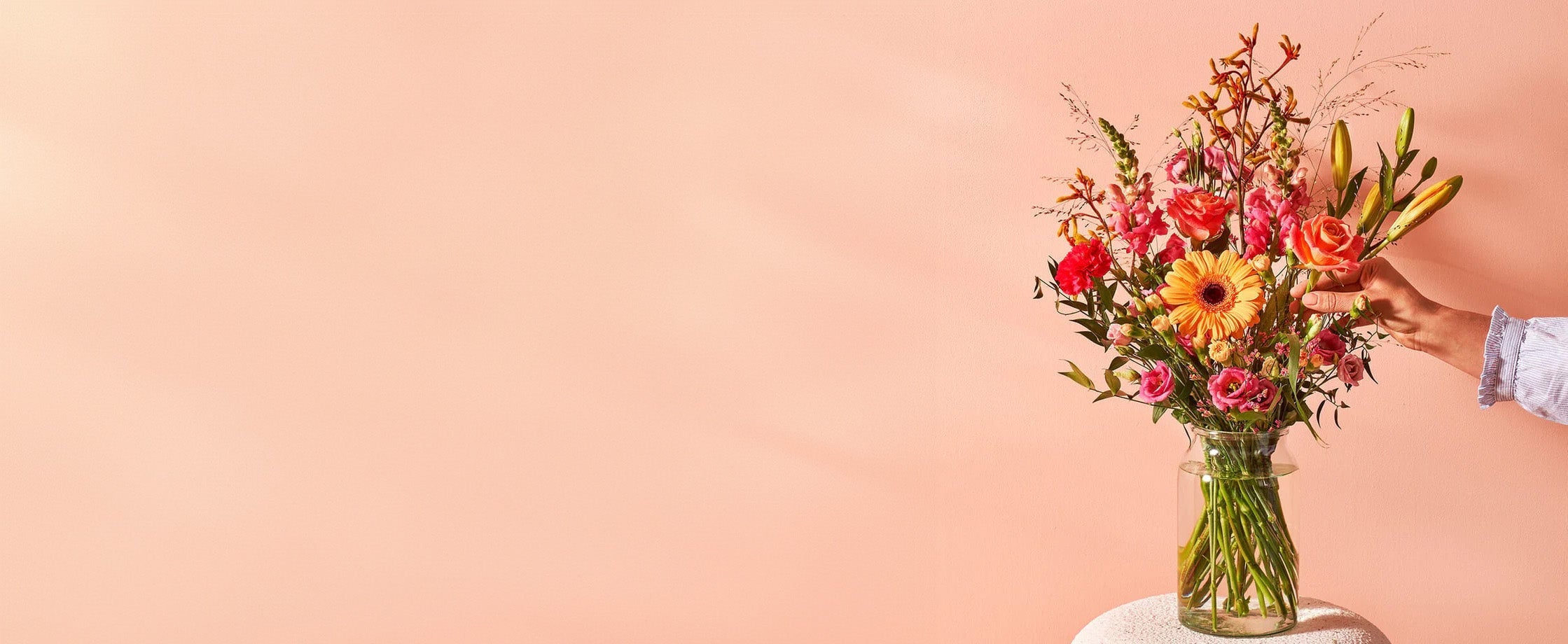 Kleurrijk boeket met lelies en gemengde bloemen in een glazen vaas tegen een lichtroze achtergrond.