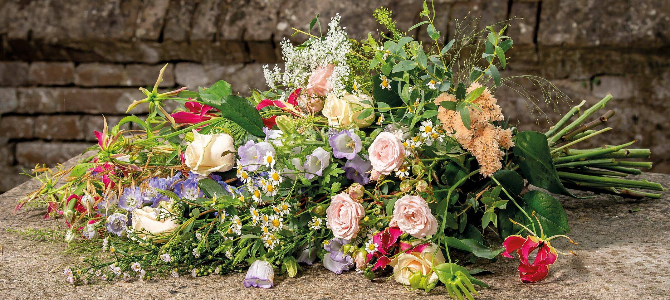 Liggend rouwboeket met natuurlijke bloemen in zachte kleuren, op een stenen achtergrond.