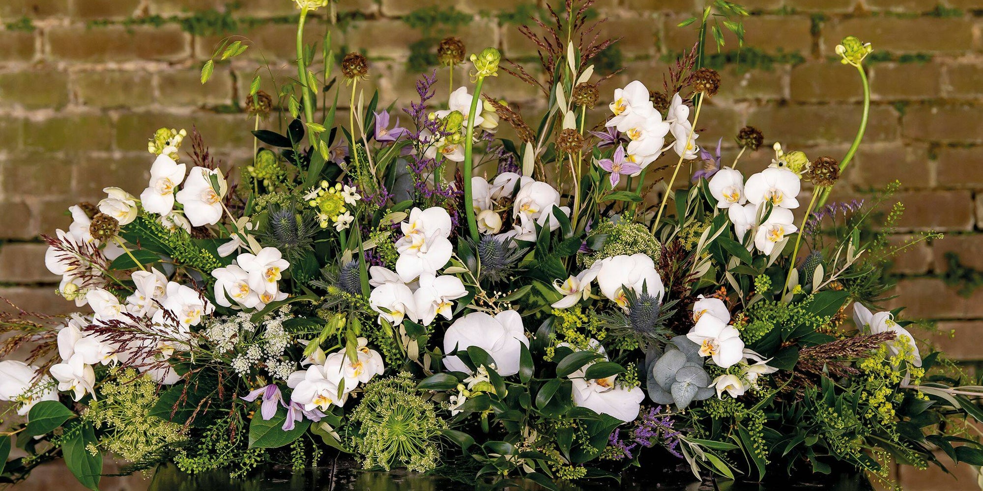 Een puur en elegant bloemstuk met witte orchideeën en subtiele paarse accenten, omlijst door weelderige groene bladeren en takken.