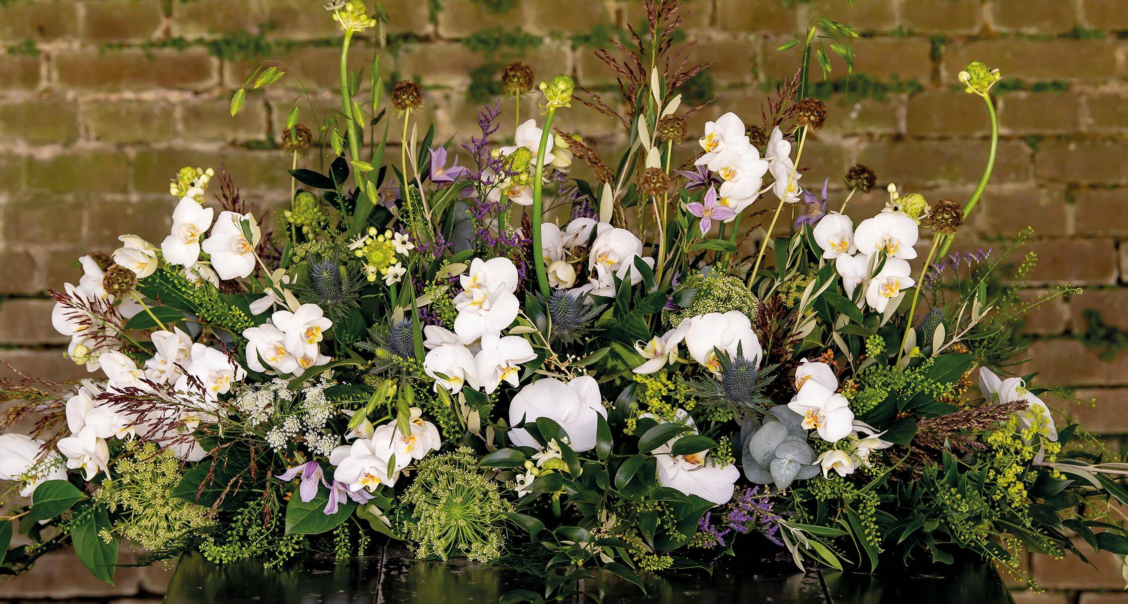 Een puur en elegant bloemstuk met witte orchideeën en subtiele paarse accenten, omlijst door weelderige groene bladeren en takken.