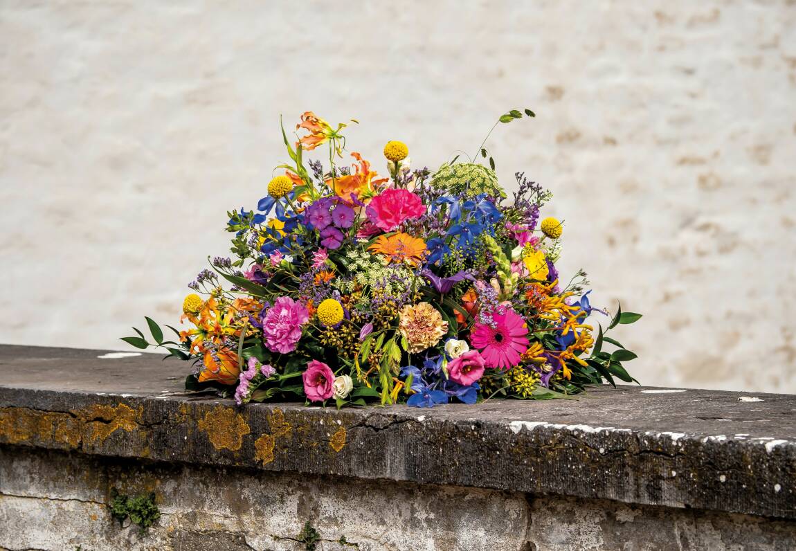 Biedermeier rouwstuk met kleurrijke bloemen, geplaatst op een stenen rand tegen een lichte achtergrond.