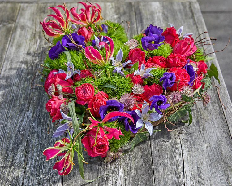 Hartvormig rouwstuk met roze en paarse bloemen op een houten ondergrond.