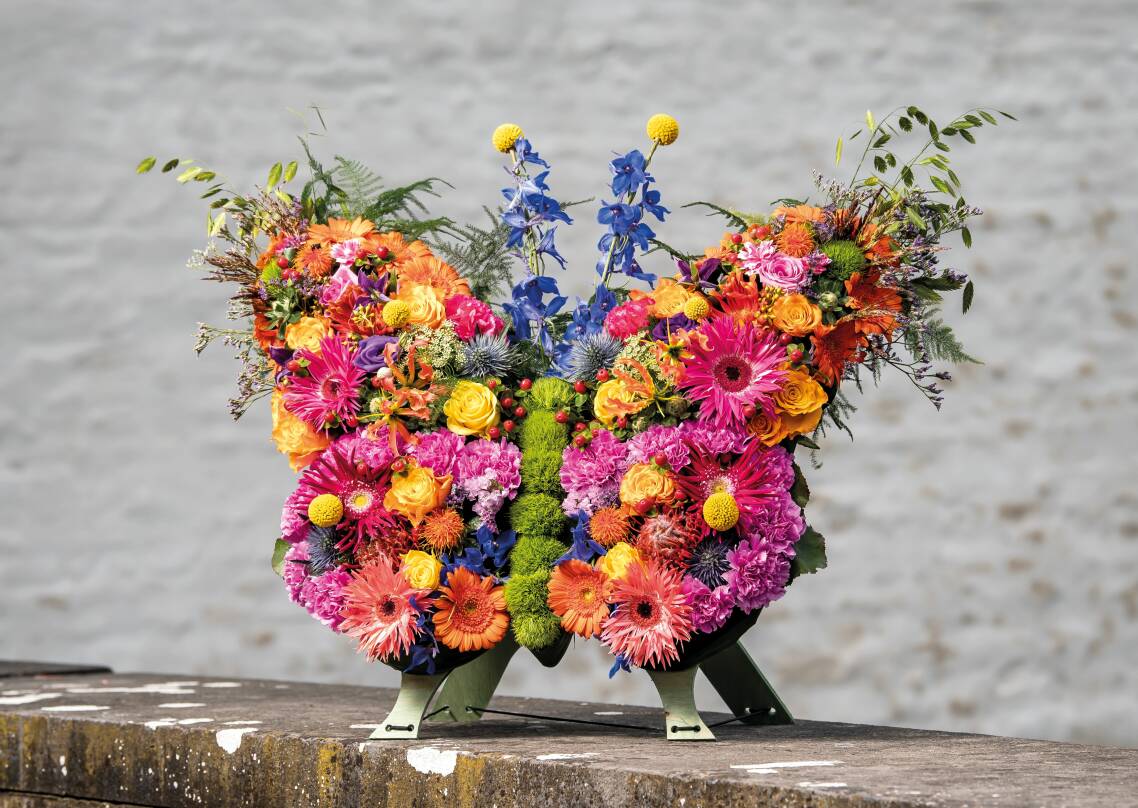 Rouwstuk in de speciale vorm van een kleurrijke vlinder, samengesteld met diverse bloemen zoals gerbera’s, rozen en chrysanten.