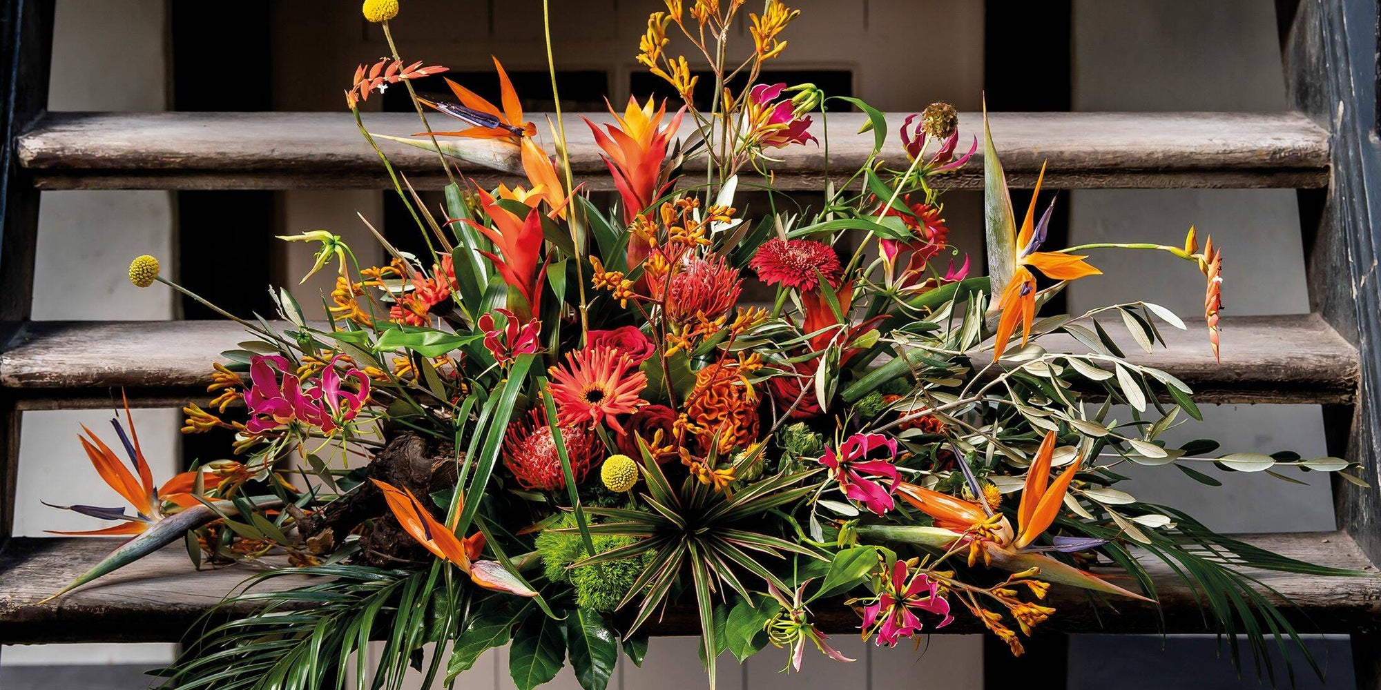 Een levendig bloemstuk met exotische bloemen in warme tinten zoals oranje en rood, gecombineerd met groene bladeren en takken, geplaatst op een houten trap.