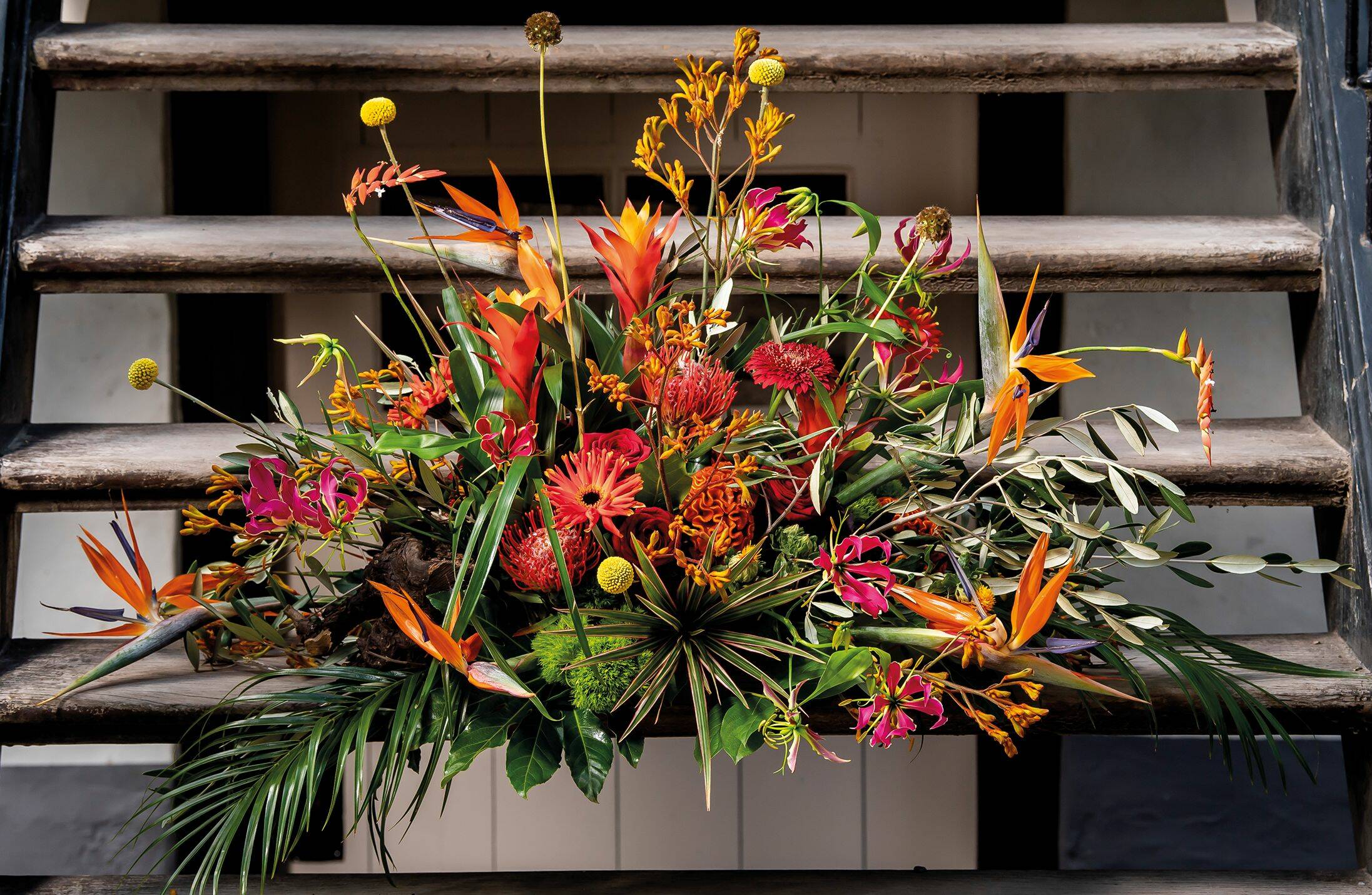 Een levendig bloemstuk met exotische bloemen in warme tinten zoals oranje en rood, gecombineerd met groene bladeren en takken, geplaatst op een houten trap.