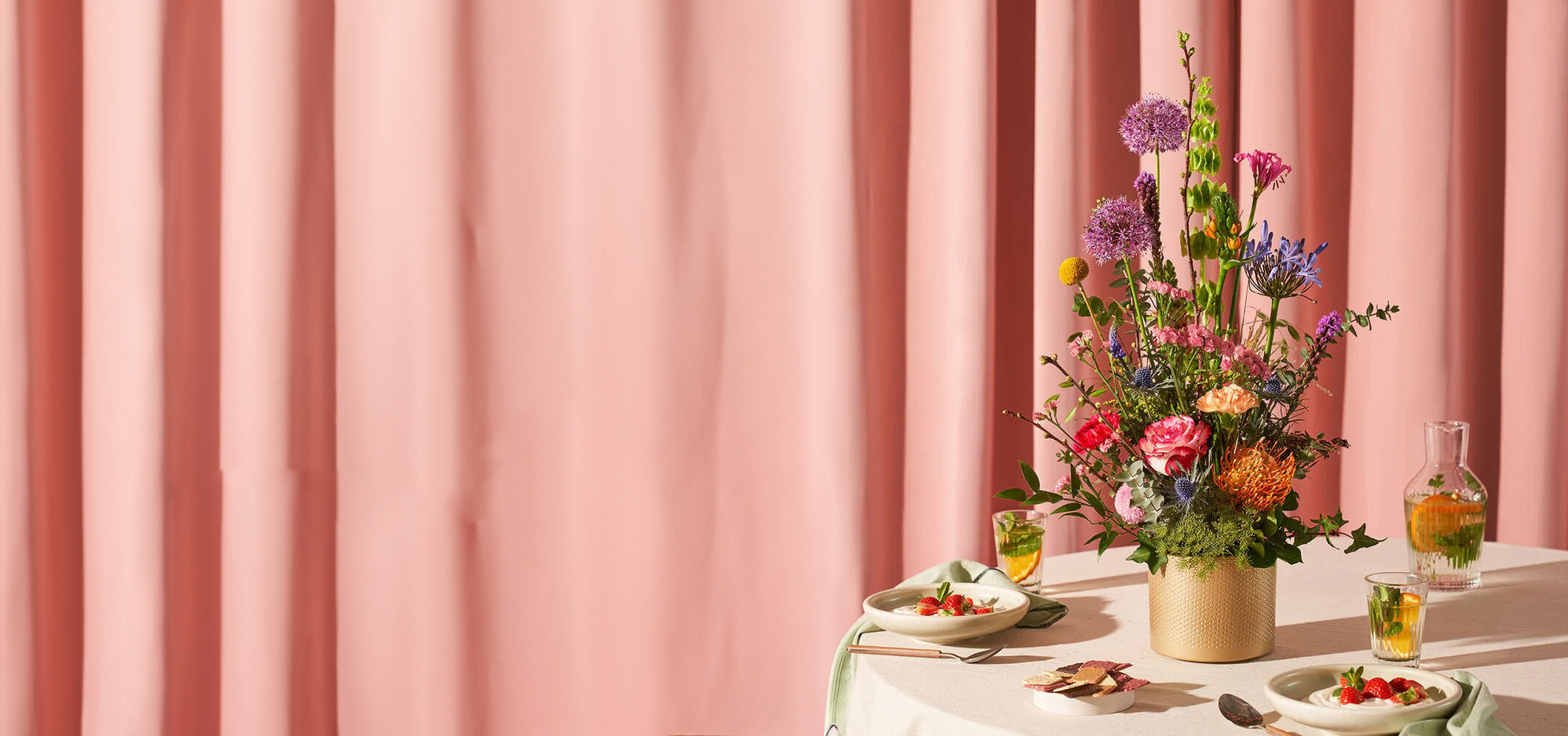 Bedankt boeket met kleurrijke bloemen in een gouden pot op een tafel gedekt met hapjes en drankjes tegen een roze gordijn