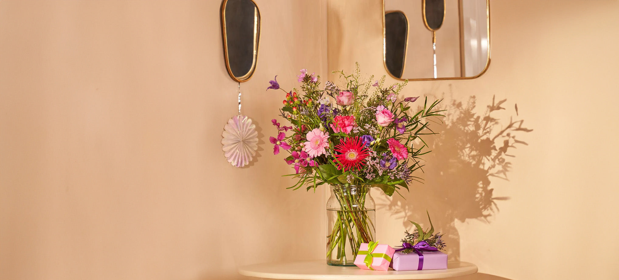 Boeket met kleurrijke bloemen in een glazen vaas naast ingepakte cadeaus, geplaatst op een ronde tafel in een huiselijke omgeving.