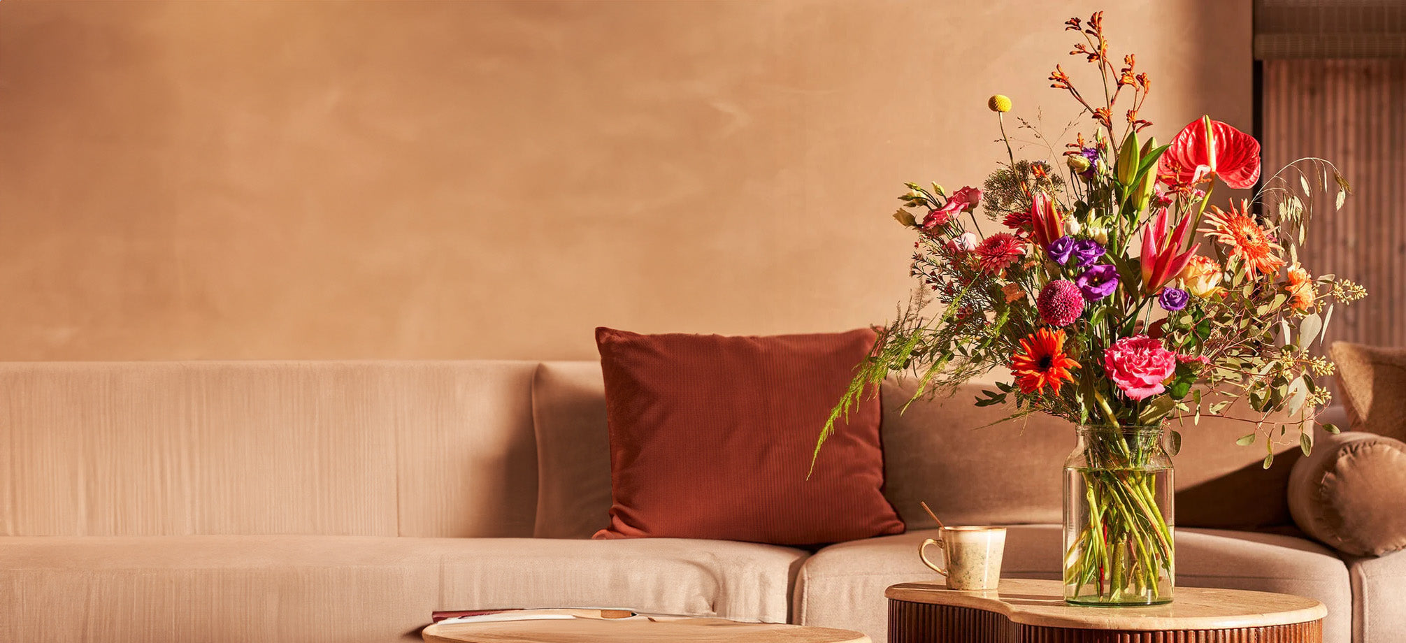 Veldboeket met kleurrijke bloemen in een glazen vaas, geplaatst op een tafel in een huiselijke setting met een beige bank.