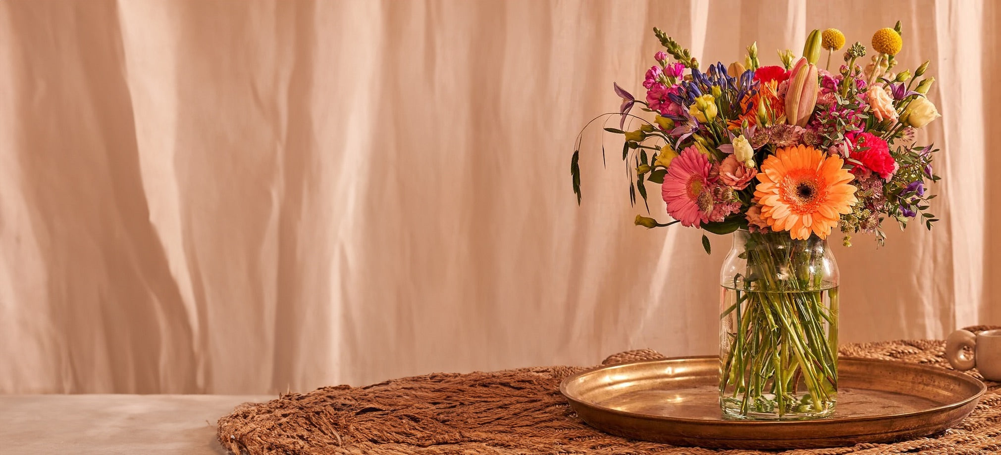 Gemengd boeket met kleurrijke bloemen in een glazen vaas op een gouden dienblad tegen een lichte achtergrond.