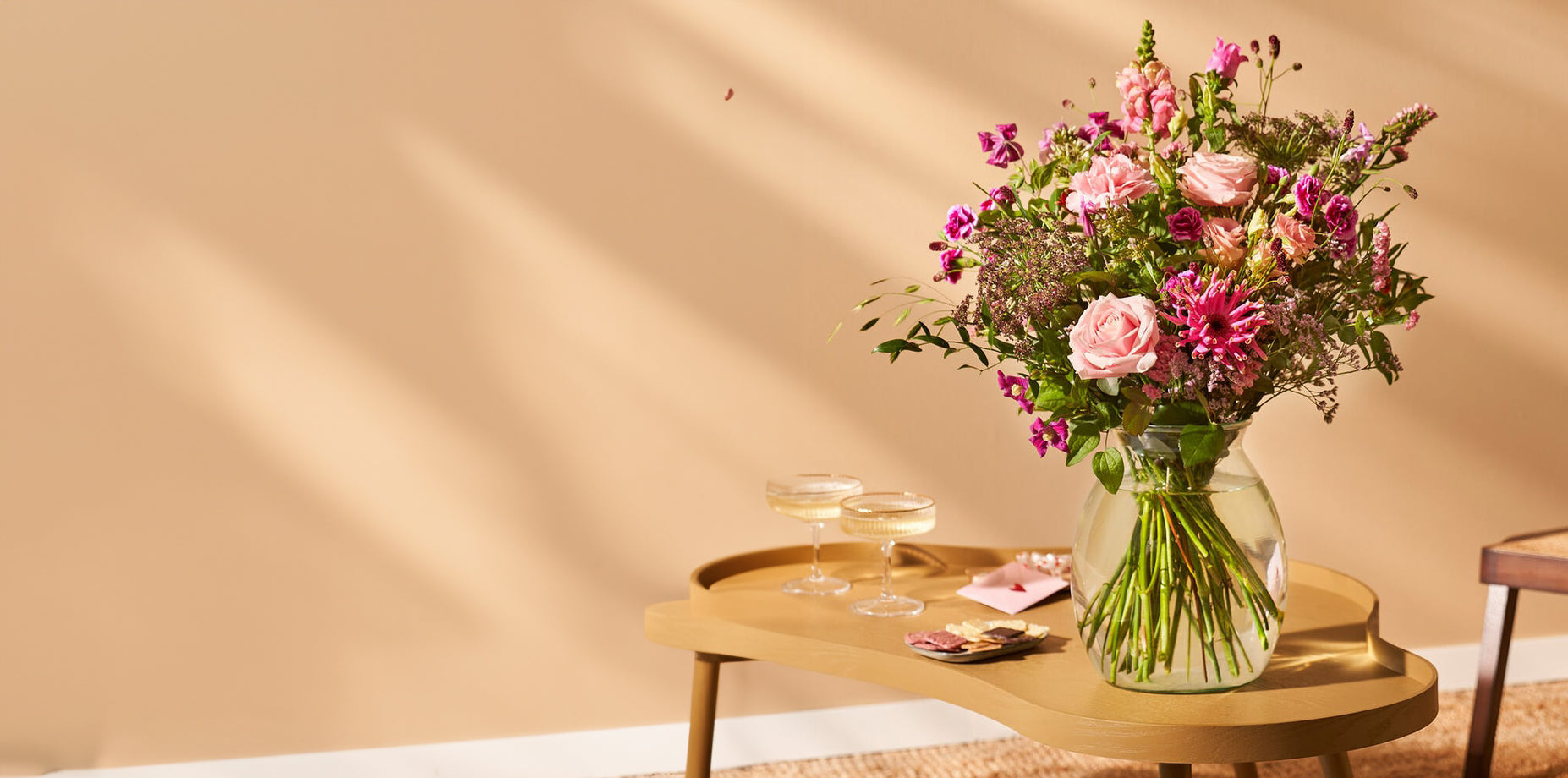 Rond boeket met roze bloemen in een glazen vaas, geplaatst op een houten tafel met champagneglazen tegen een beige achtergrond.