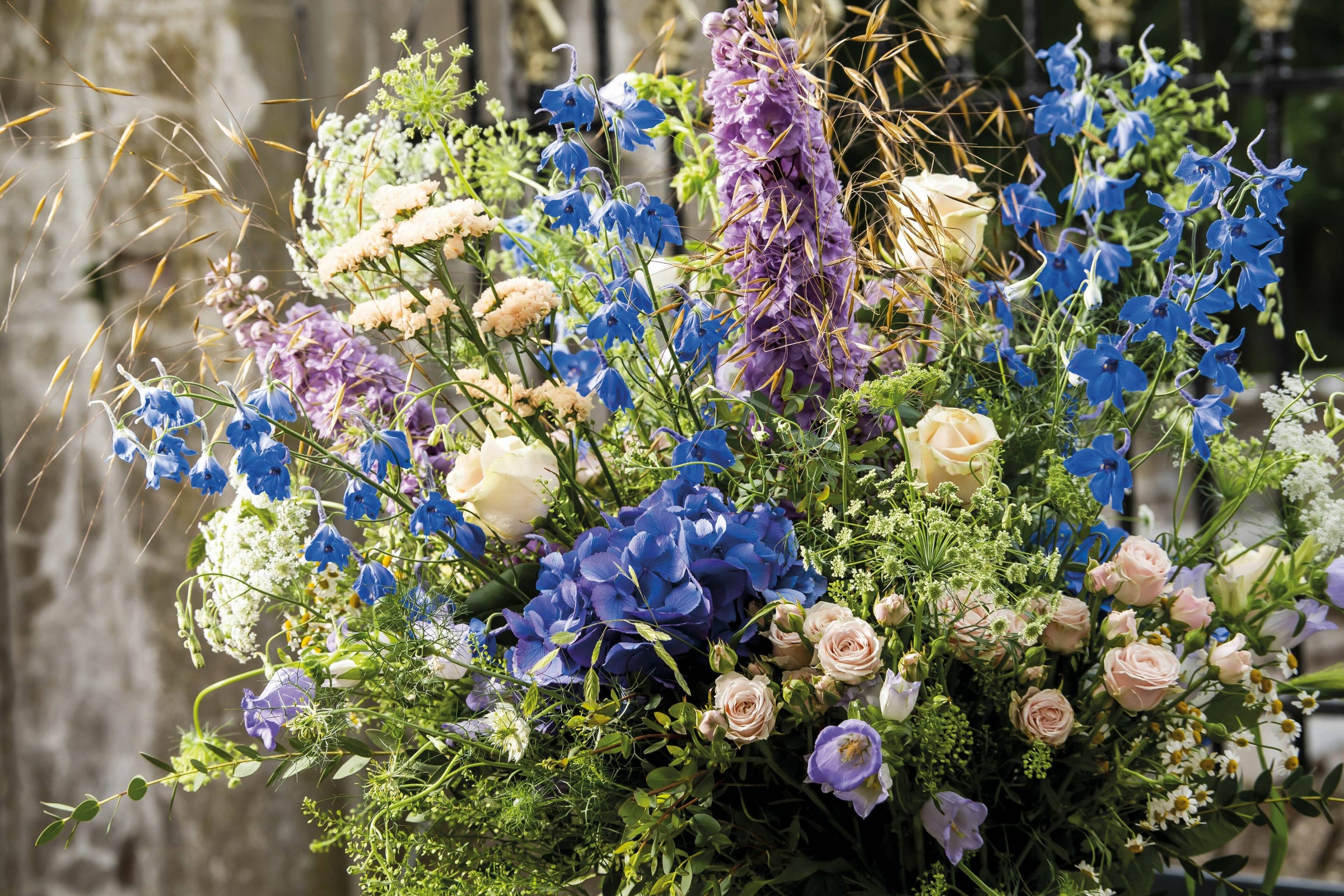 Rouwbloemen arrangement met een mix van blauwe delphiniums, paarse hortensia’s, rozen en seizoensbloemen in zachte tinten.