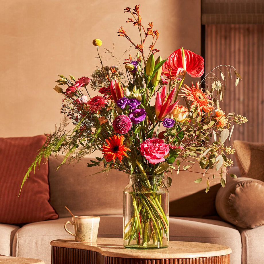 Kleurrijk boeket in een glazen vaas met anthurium, lelie, roos en andere bloemen, op een bijzettafel in een gezellige woonkamer setting.