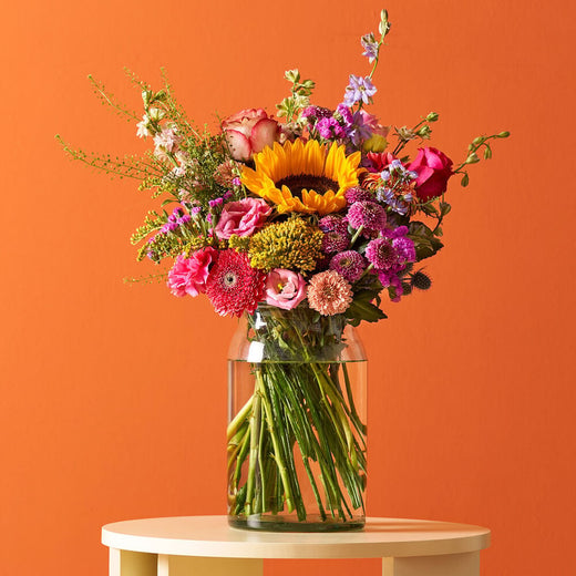 Kleurrijk boeket in een glazen vaas met zonnebloemen, rozen en andere bloemen, op een witte tafel tegen een oranje achtergrond.