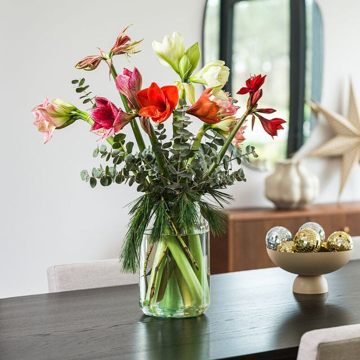 Boeket met kleurrijke amaryllis bloemen en dennengroen in glazen vaas, gestyled in een modern interieur.