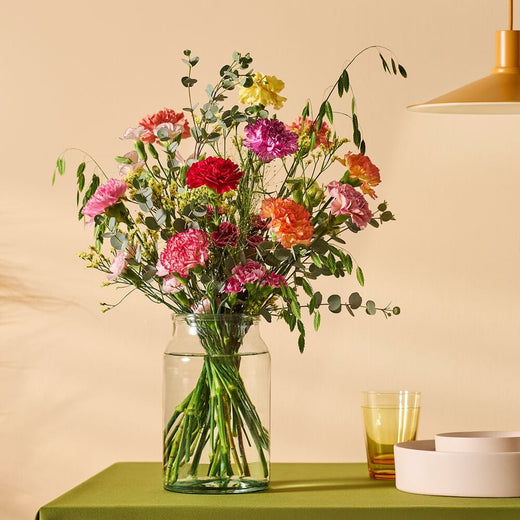 Boeket met kleurrijke anjers in een glazen vaas, gestyled op een tafel met groen tafelkleed en moderne decoratie.