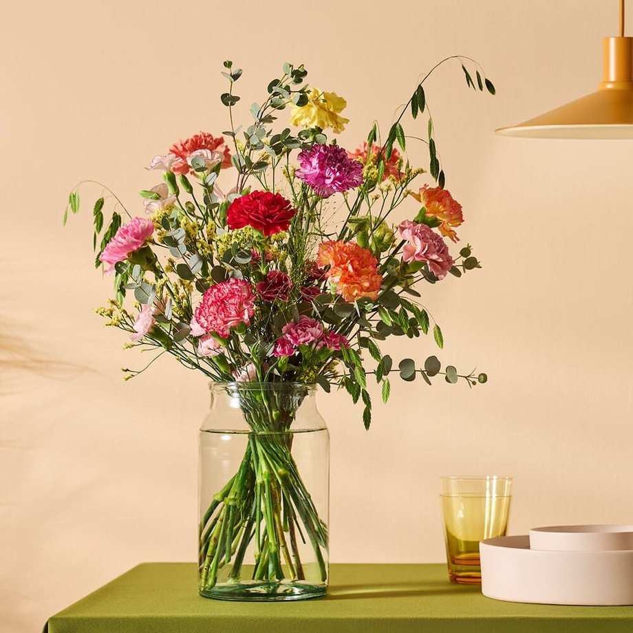 Boeket met kleurrijke anjers in een glazen vaas, gestyled op een tafel met groen tafelkleed en moderne decoratie.
