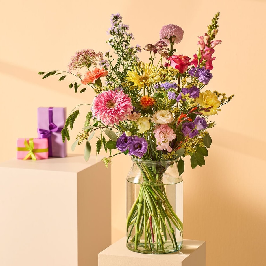 Vrolijk boeket met kleurrijke bloemen zoals gerbera’s en lisianthus in een glazen vaas, tegen een gele achtergrond.