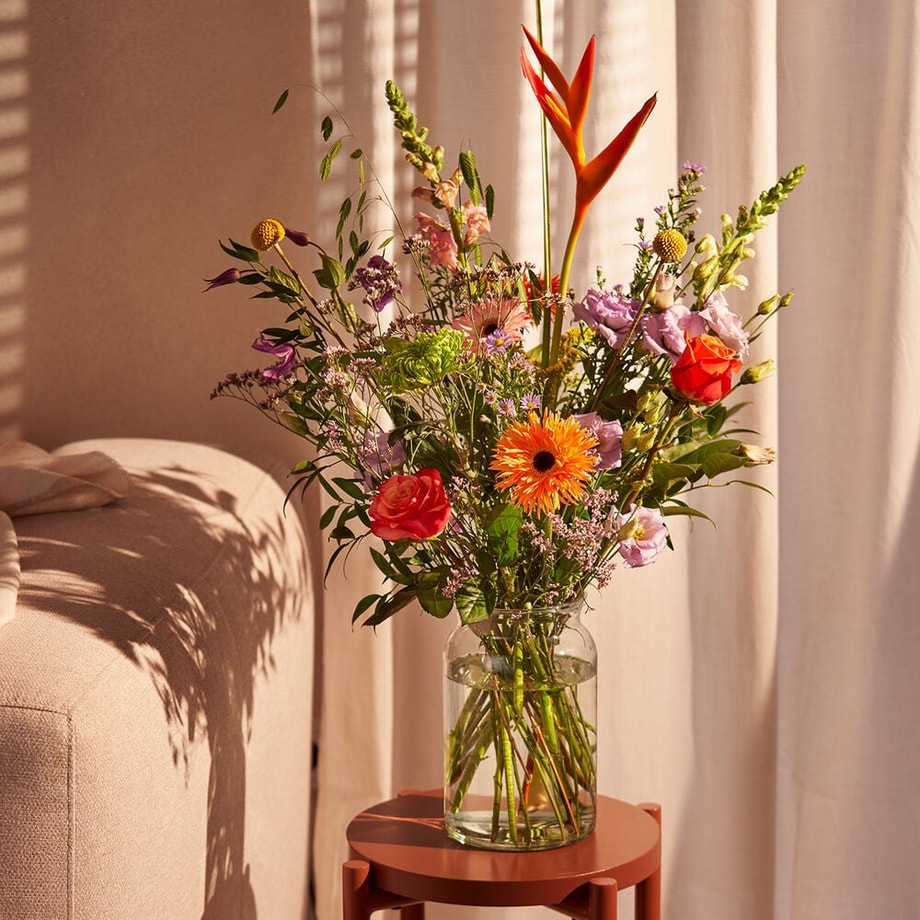 Kleurrijk boeket in een glazen vaas met paradijsvogelbloemen, rozen en gerbera's, op een tafel in een lichte woonkamer setting.