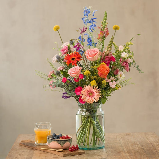 Bont boeket met roze, oranje en blauwe bloemen, waaronder rozen en gerbera’s, in glazen vaas op houten tafel met ontbijt en sinaasappelsap
