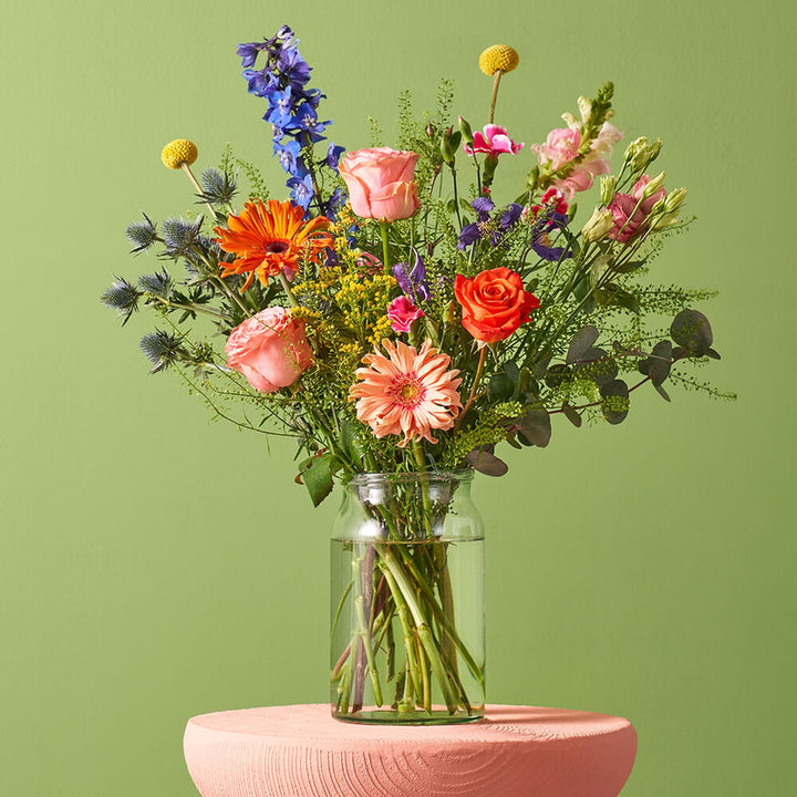 Kleurrijk boeket in een glazen vaas met delphinium, rozen, gerbera's en andere bloemen, op een roze tafel tegen een groene achtergrond.