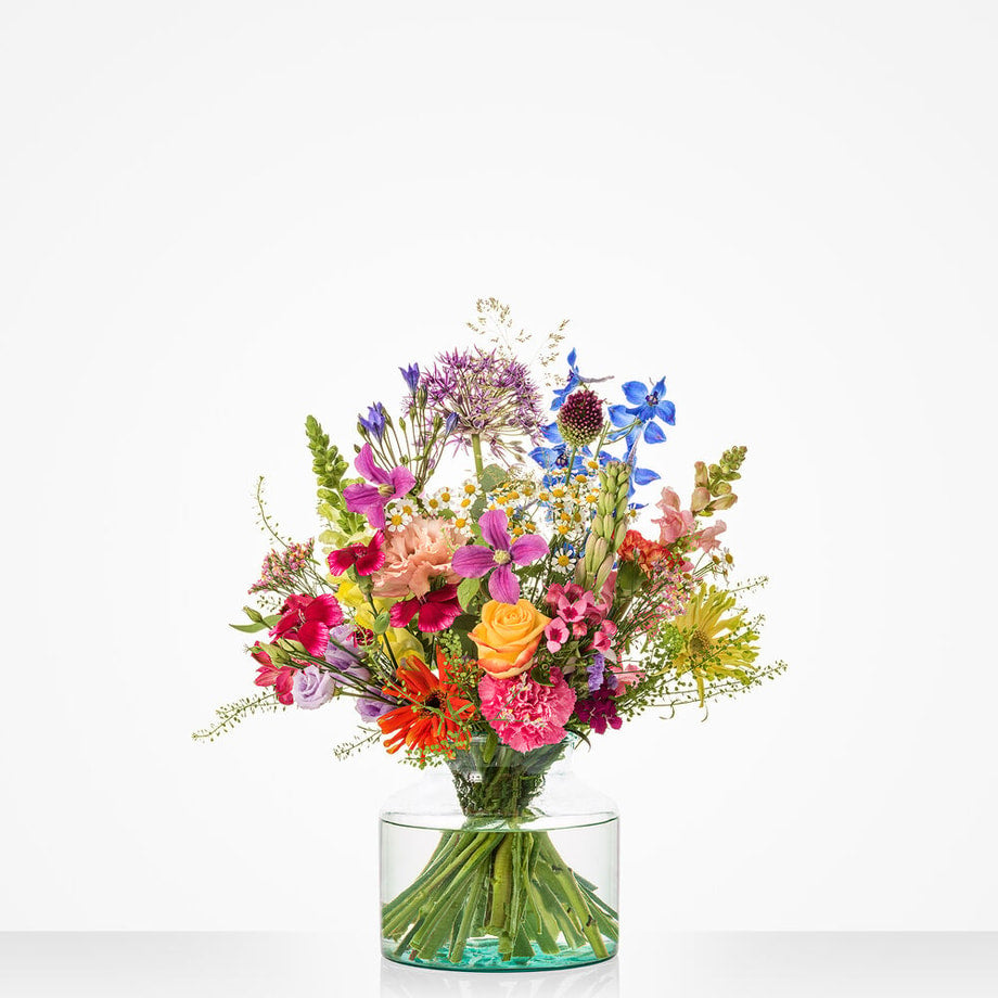Groot veldboeket met kleurrijke bloemen in tinten van geel, oranje, roze, paars en blauw in een glazen vaas