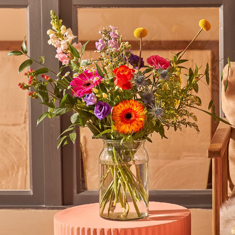 Bont boeket met oranje, roze en paarse bloemen, waaronder gerbera’s en rozen, in glazen vaas op roze tafel in zonlicht