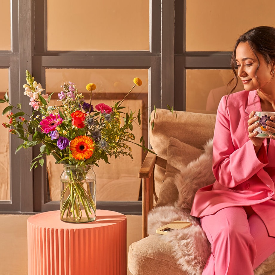 Bont gekleurd boeket in een glazen vaas met gerbera's, rozen en andere bloemen, op een roze tafel in een gezellige kamer met een vrouw in een roze pak.