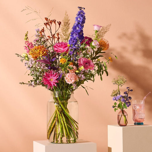 Deluxe veldboeket met kleurrijke bloemen in glazen vaas, sfeervol gepresenteerd tegen een perzikkleurige achtergrond met kleine bijpassende vaasjes en een roze drankje