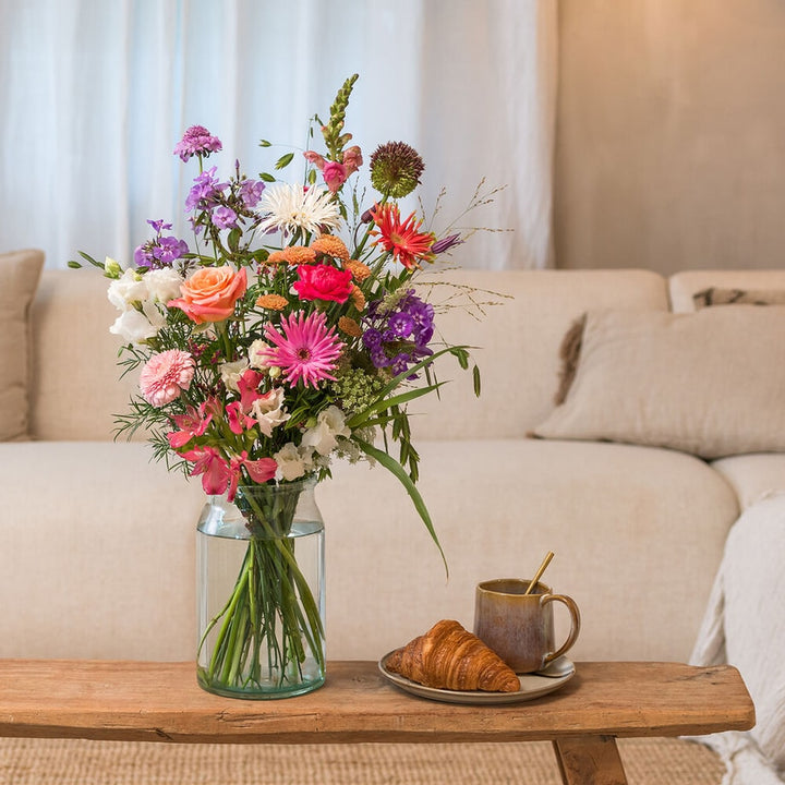 Kleurrijk boeket met roze, paarse en oranje bloemen in glazen vaas op houten tafel met croissant en koffie