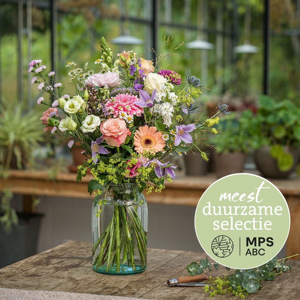 Duurzaam pastelboeket met rozen, gerbera’s en veldbloemen in glazen vaas in kasomgeving