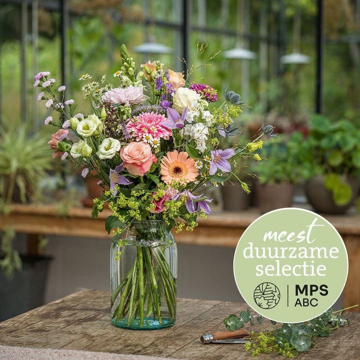 Duurzaam pastelboeket met rozen, gerbera’s en veldbloemen in glazen vaas in kasomgeving